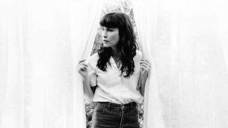 "A black-and-white photo of a woman (Missy Higgins) with wavy hair standing between two parted sheer curtains. She is wearing a white button-up blouse tucked into high-waisted jeans and looks thoughtfully to the right, with a calm and contemplative expression. The soft lighting and minimal background create an introspective mood.