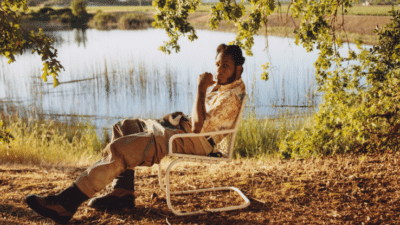 A man with short hair and a goatee sits on a white folding chair under the shade of a tree, overlooking a peaceful lake. He is wearing a patterned short-sleeve shirt, loose beige pants, and black boots. The natural setting includes a calm body of water with reeds and a distant green landscape under soft, natural light.