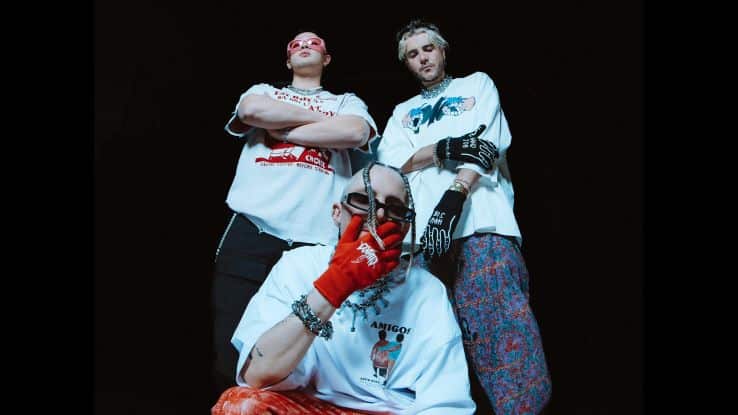 The image shows three people standing in a bold and confident pose against a dark background. They are dressed in streetwear-style clothing, with oversized graphic T-shirts, patterned pants, and accessorized with chains, rings, and gloves. One individual in the front is wearing red gloves, sunglasses, and has their hand raised in a gesture, while the others stand slightly behind with crossed arms. The overall aesthetic is edgy and urban, emphasizing a rebellious, alternative vibe.