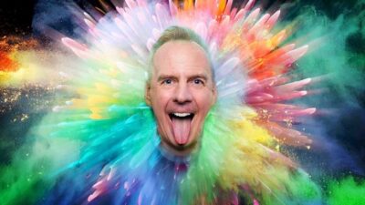 A man with a joyful expression and his tongue sticking out, set against a vibrant, multicolored explosion of paint-like splashes radiating outward. The colors range from bright greens, blues, pinks, and yellows, creating a dynamic and energetic background.