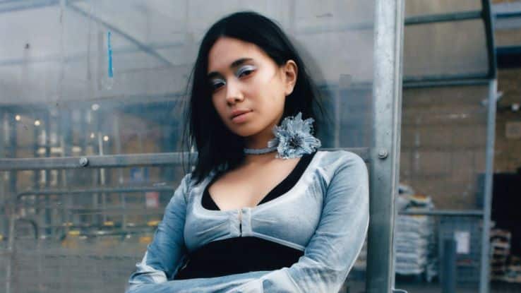 A young woman with shoulder-length dark hair sits against a metal structure in an industrial setting. She is wearing a silver-gray, long-sleeved top over a black outfit, with a large silver flower accessory on her choker and subtle white and silver eye makeup. She gazes directly at the camera with a calm expression, framed by the muted, metallic surroundings.